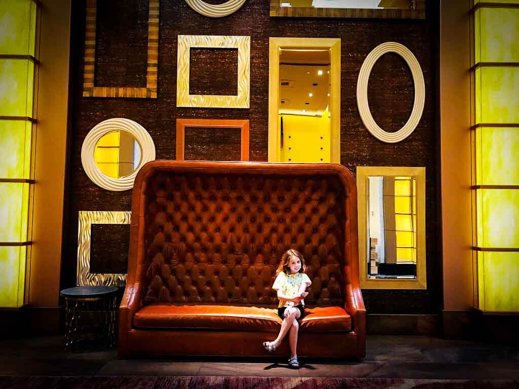 girl in tshirt and shorts sat on a high backed sofa with mirrors of all shapes on the wall behind