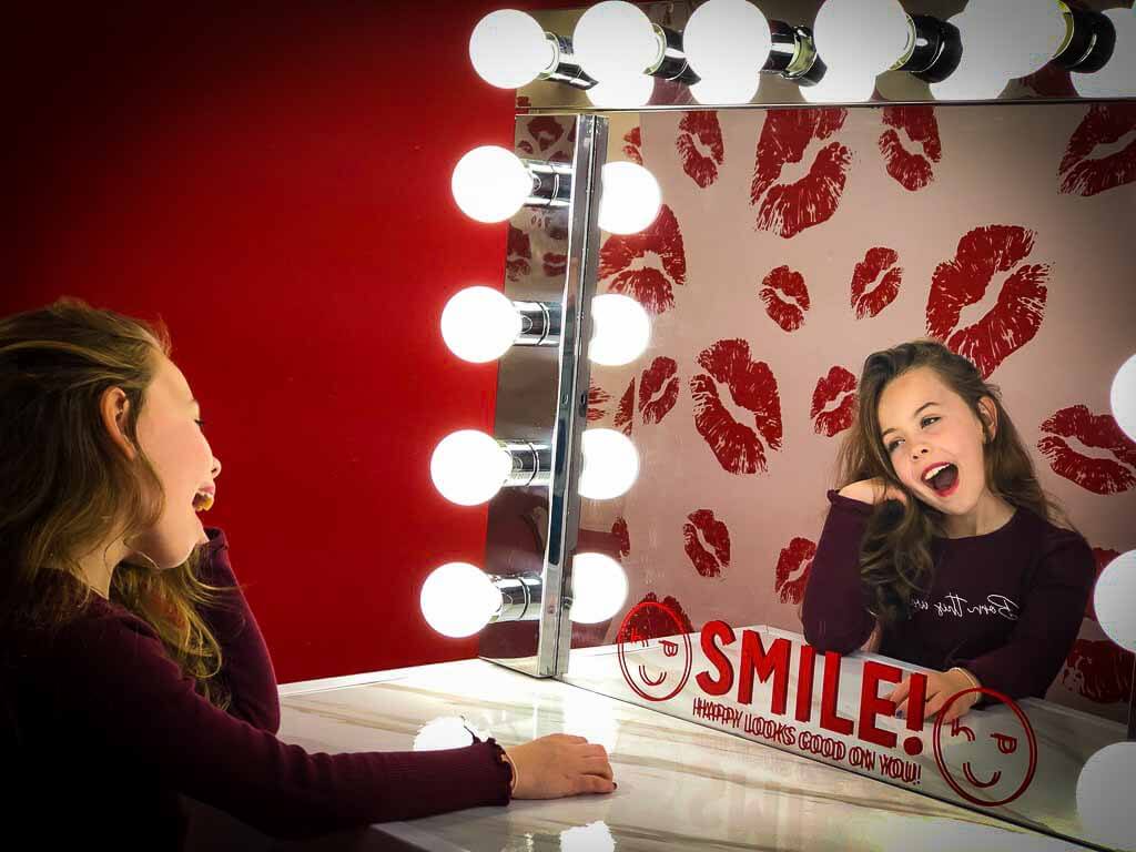 girl in front of a mirror with light bulbs around it, the mirror says smile